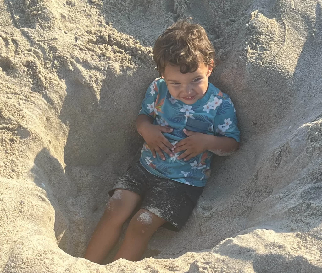 young boy laying in a big hole in the sand on beach