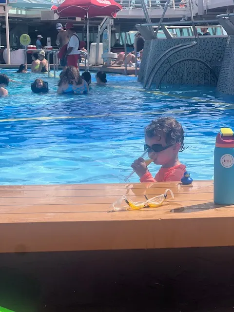 A boy toddler wearing sunglasses drinking juice in the pool.