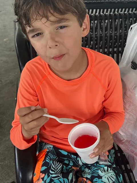 A little boy in eating a cherry snow cone sitting on bench.