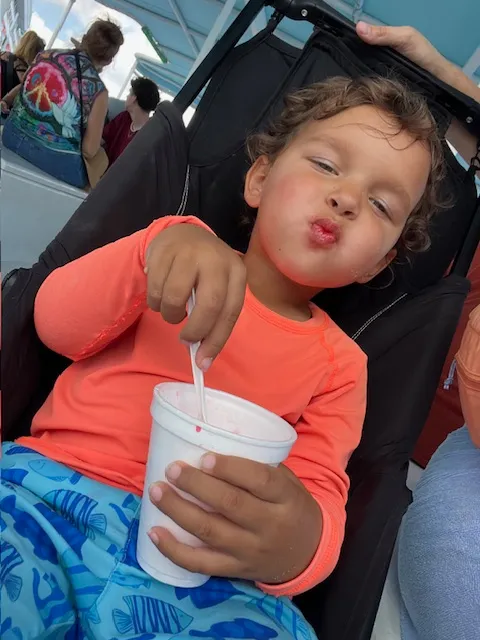 A toddler in summer clothes eating a snow cone in stroller.