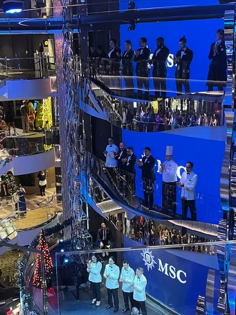 A group of chefs and crew members stand on multiple levels of a modern cruise ship's atrium, with a blue backdrop displaying "MSC." 