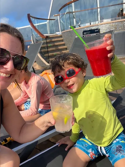 A woman and a child in sunglasses are sitting on lounge chairs, smiling and holding drinks on cruise ship deck.