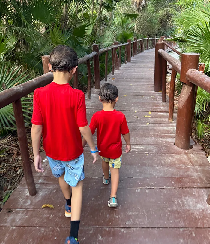 Two young children walking on a wooden bridge surrounded by lush green foliage.