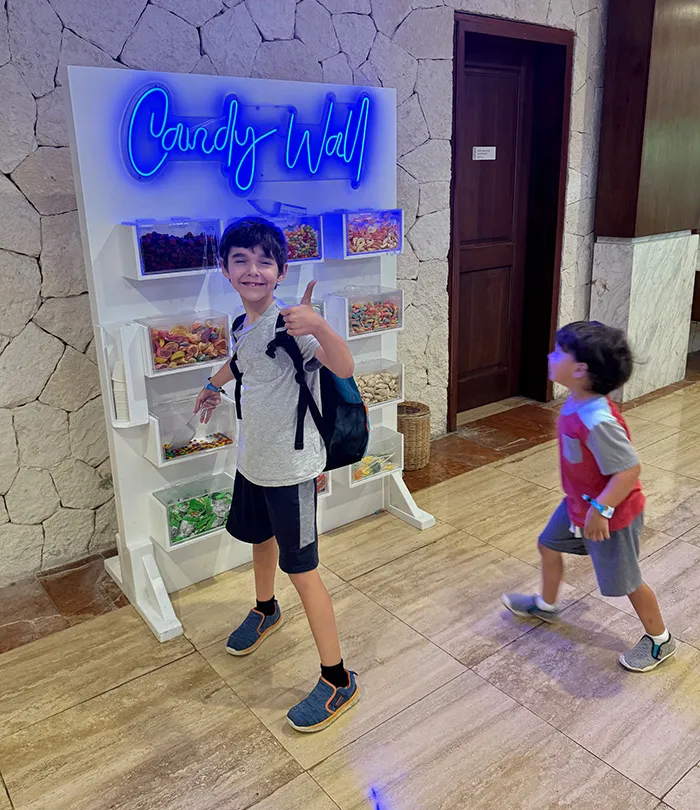Two young boys stand near a wall display labeled “Candy Wall” with various bins of colorful candy.
