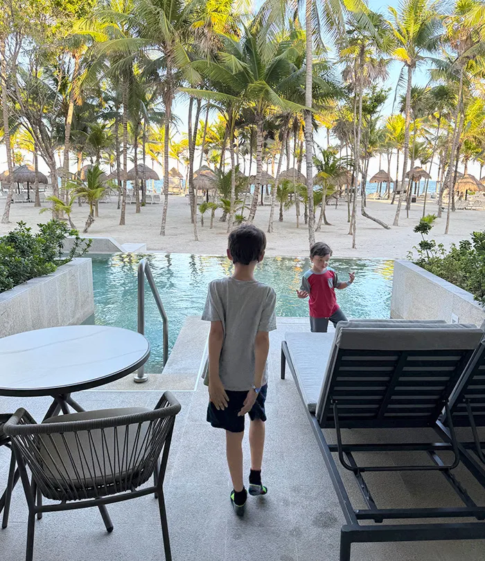 Two young boys stand by a private pool on a patio with lounge chairs and a table, overlooking a tropical beach lined with palm trees and thatched umbrellas.