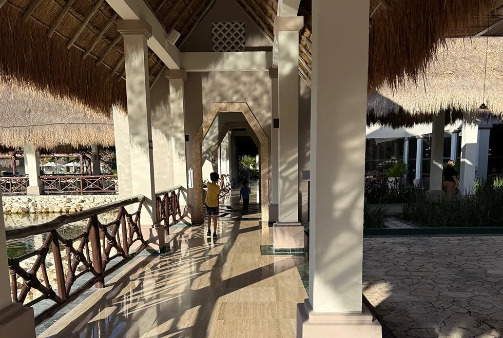 A sunlit, covered walkway with a thatched roof, flanked by wooden railings and columns with 2 children walking.