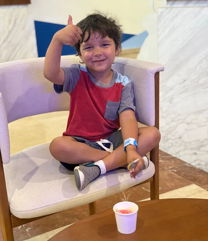 A young boy with sitting cross-legged on a chair, smiling and giving a thumbs up.