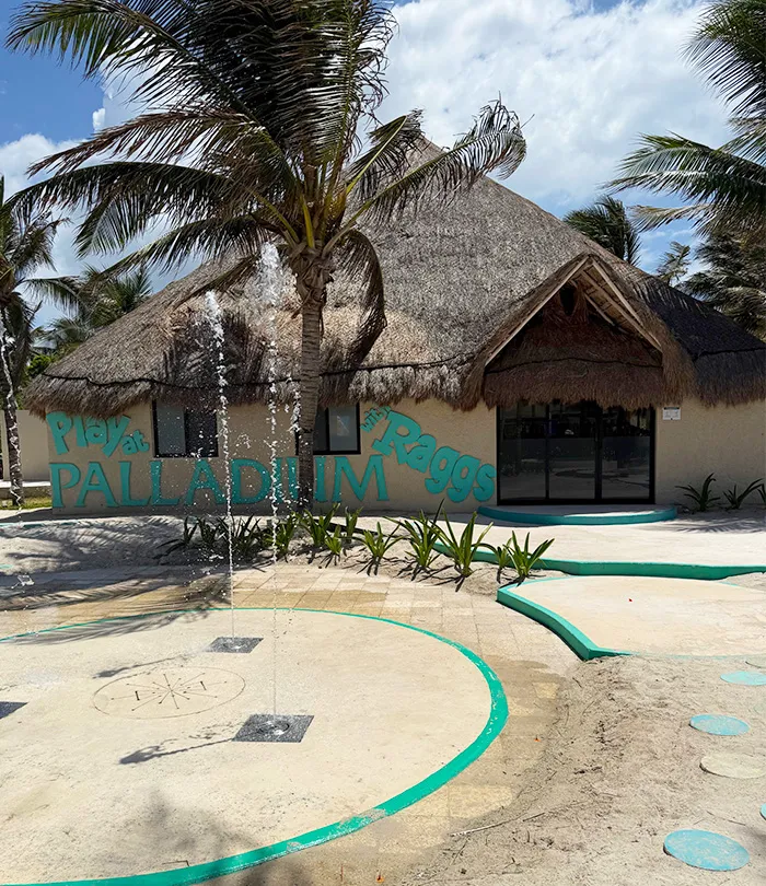 A thatched-roof building with signs reading "Playa Palladium" and "Raggs" is surrounded by palm trees. A small water fountain sprays jets of water in front of the entrance under a partly cloudy sky.