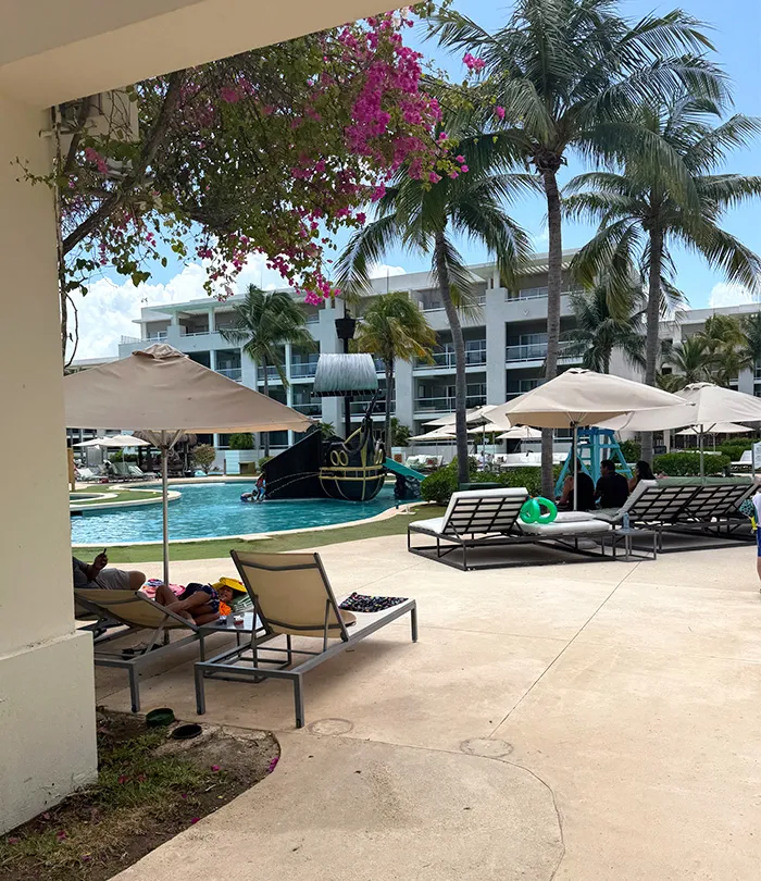 Poolside scene at a resort with lounge chairs, palm trees, and a pirate ship-themed play structure in the pool.