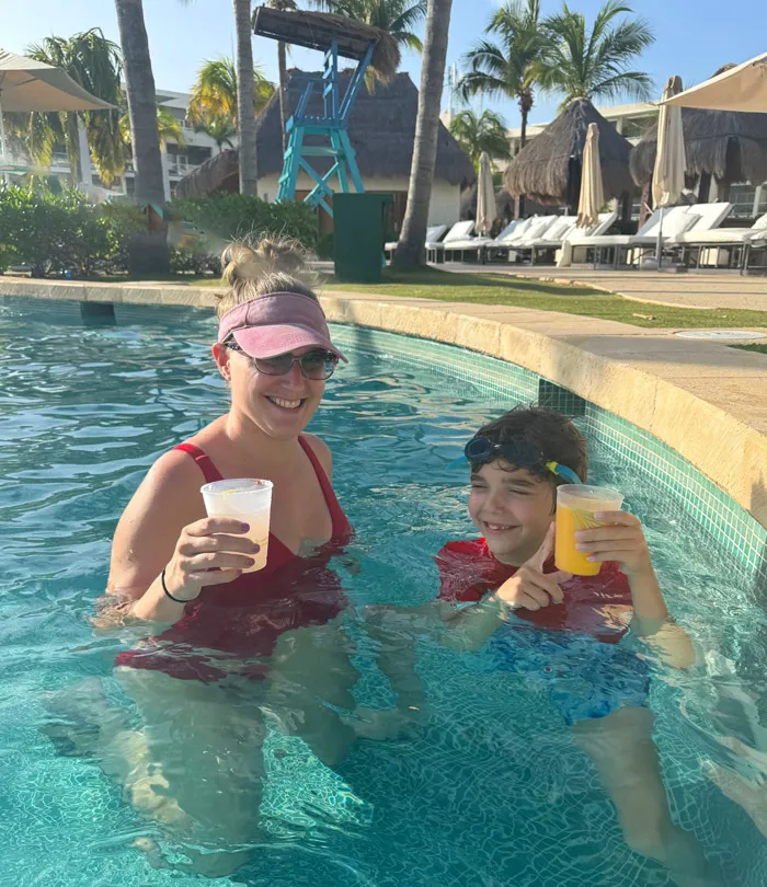 A mother and child smile and are holding drinks while in an outdoor pool.