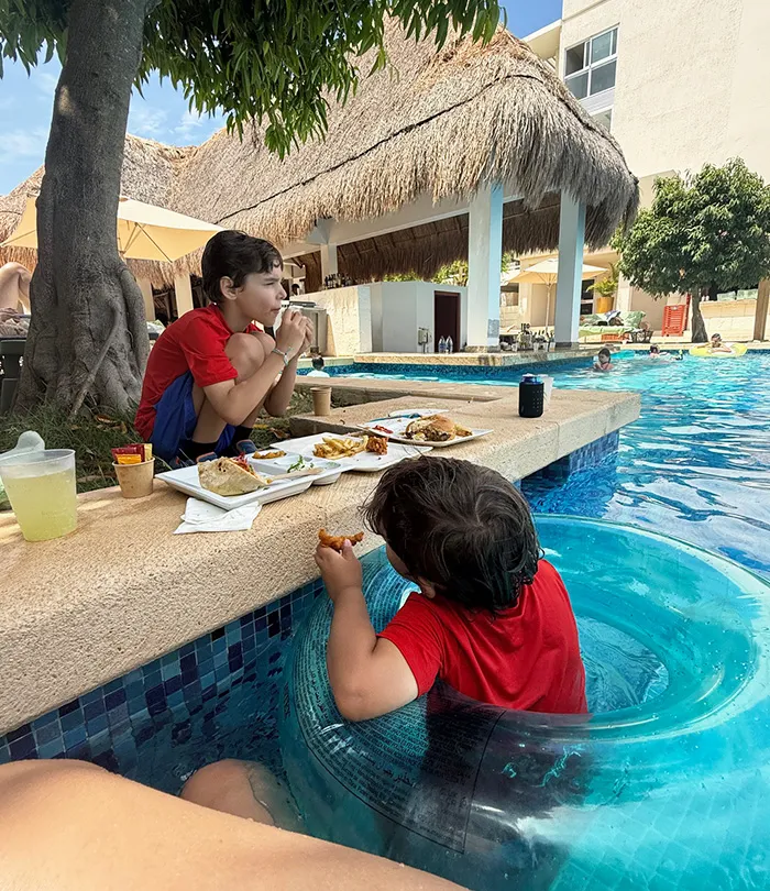Two young boys in red shirts eat food by a pool, with one sitting at the edge, while the other sits in the water.