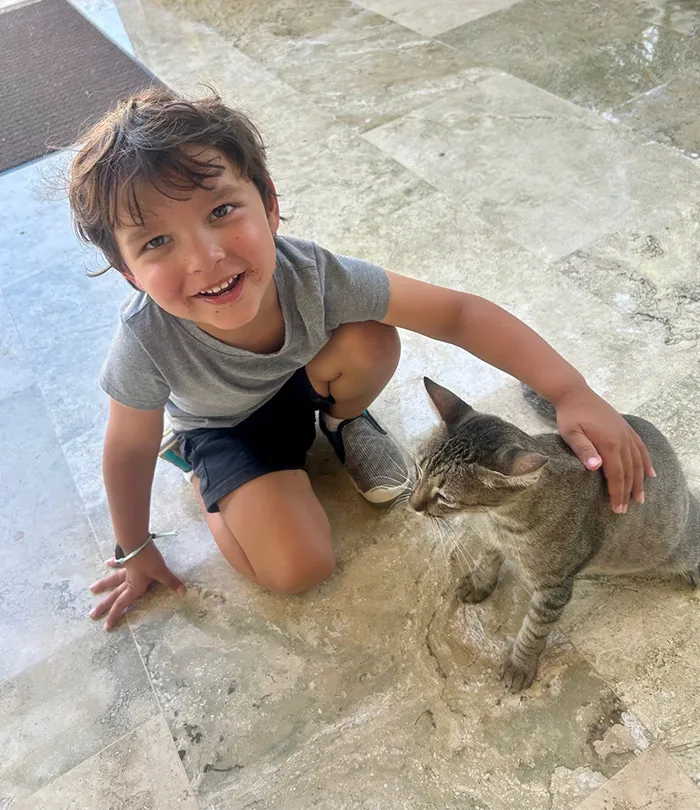 A young boy kneeling with brown hair, wearing a t-shirt and shorts, petting a gray tabby cat beside him.