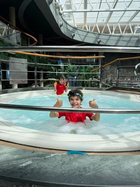 Two young boys in red swim shirts swimming and giving thumbs up in an MSC cruise ship pool area.