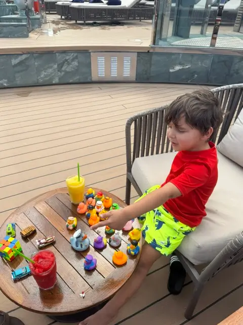 A young boy playing with cruise ducks, toy cars, and colorful frozen drinks on a outdoor deck table.
