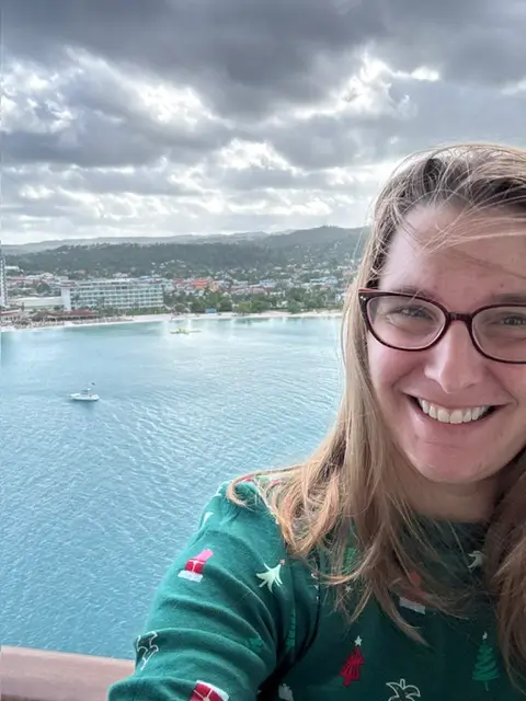 Sara, owner of The Traveling Mommune, smiling in a festive green sweatshirt on an MSC Seashore balcony overlooking a Caribbean port.
