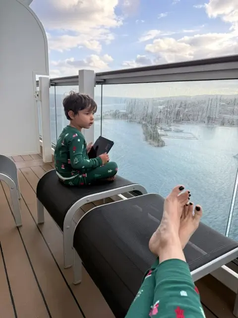 A young boy sitting on a lounge chair wearing Christmas pajamas on a private cruise balcony overlooking the ocean.