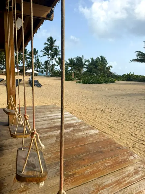 Wooden beach bar swings on a deck overlooking the quiet, palm-lined beach.